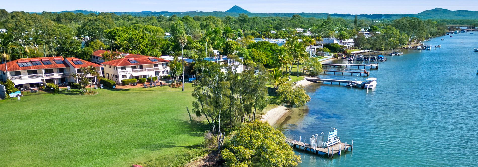 Aerial view of Cayman Quays Noosaville holiday property highlighting the private jetty, resort facade, expansive Noosa River, and distant hinterland.
