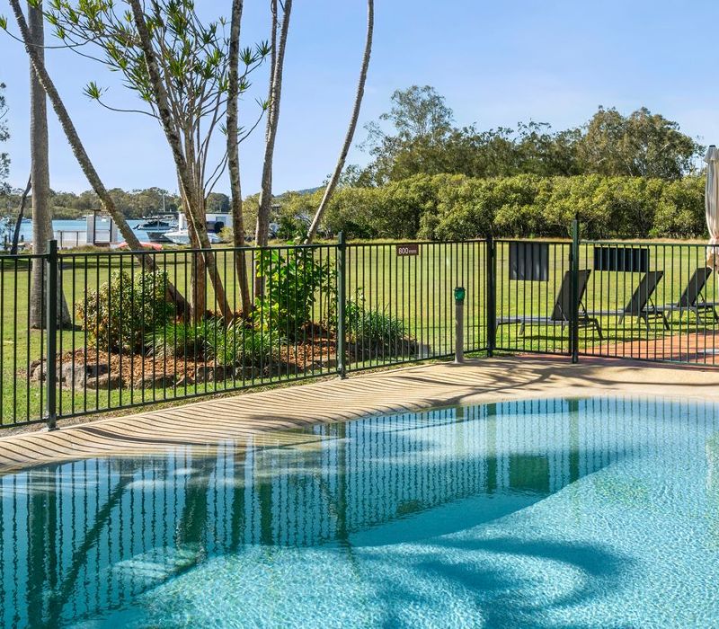 Panoramic view of the resort swimming pool featuring chaise sun lounges arranged outside the safety gate with the Noosa River in the background.
