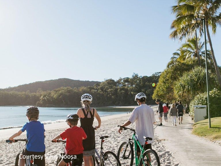 A family riding bicycles along the flat, scenic boardwalk of Noosa Main Beach, using complimentary guest gear from Cayman Quays.