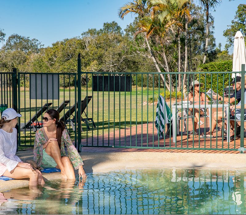 Mother and daughter sitting on the pool edge while a couple relaxes at an outdoor dining table adjacent to the green parkland leading down to the Noosa River.