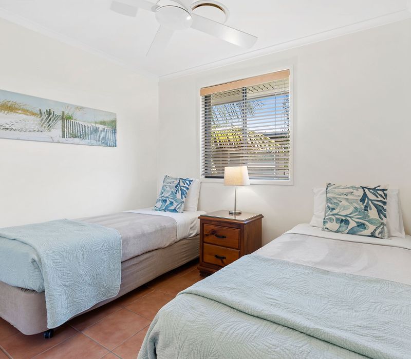 Spacious holiday apartment bedroom styled with one twin single bed and one double bed beneath large windows framing regional landscape views.