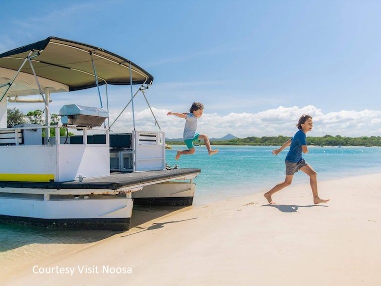 Children enjoying watersports and jumping from a hired flat-boat catamaran directly onto a sandy beach along the Noosa River.