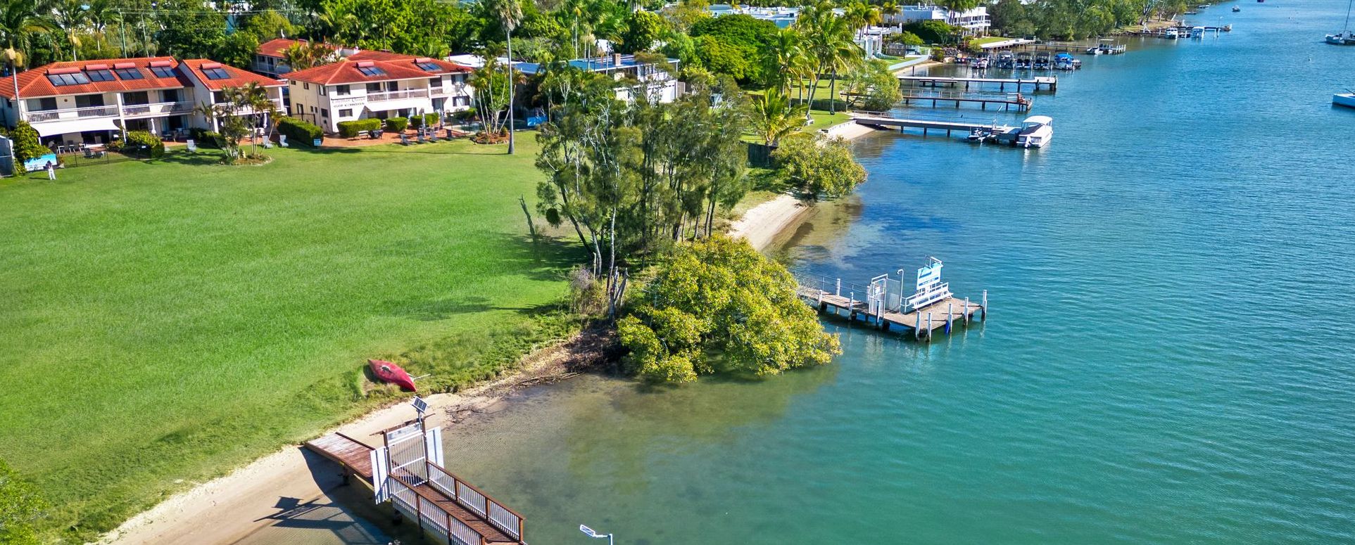 High-angle aerial capture of Cayman Quays Noosa Riverfront resort highlighting the private guest jetty, expansive manicured grass lawn, and panoramic views of the water.