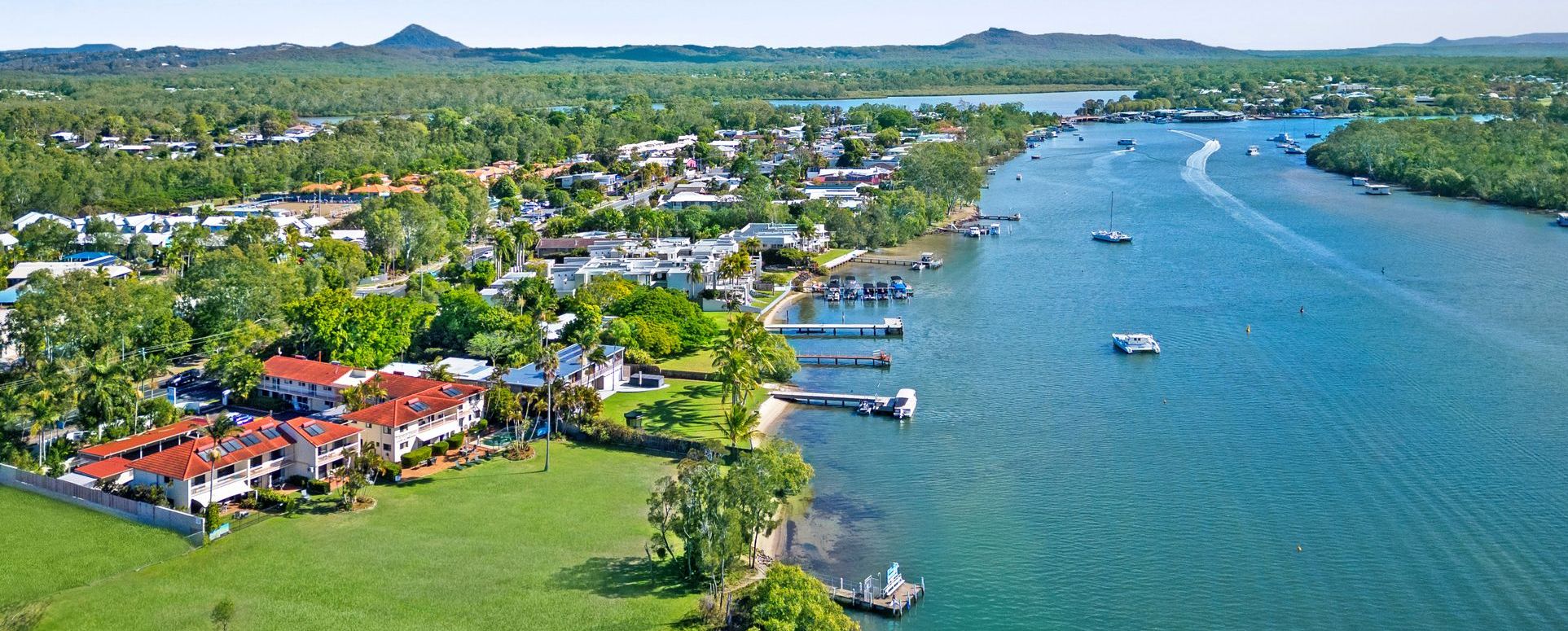 Aerial view of Cayman Quays holiday apartments in Noosaville showing the 70m absolute riverfront grassland leading to the Noosa River, private guest boat mooring, and distant Sunshine Coast hinterland mountains.