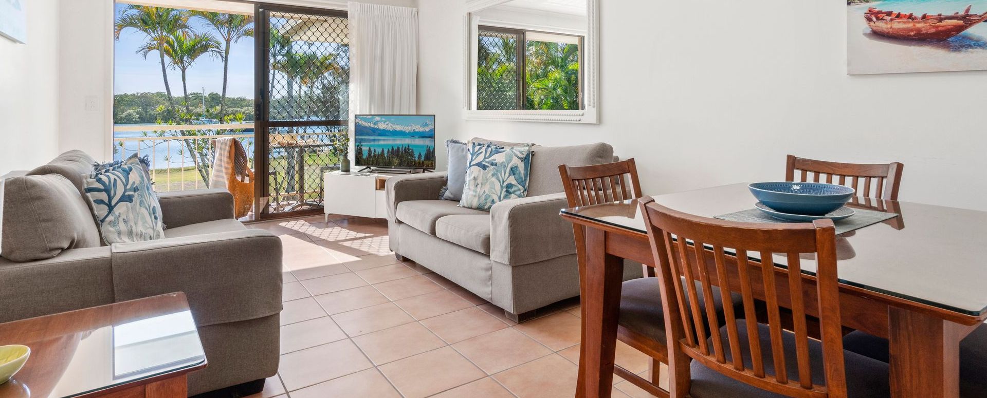 Comfortably furnished apartment living room with a large flat-screen TV and dining table, looking out through glass sliders toward the private patio and the Noosa River.