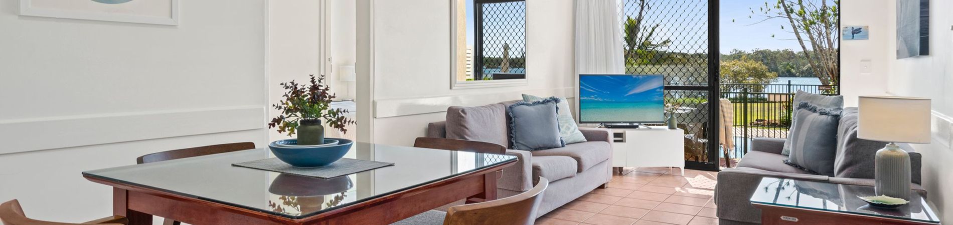 Wide open-plan view of the dining area and lounge room with a big screen TV, featuring glass sliding doors that open out to a private balcony with direct Noosa River views.