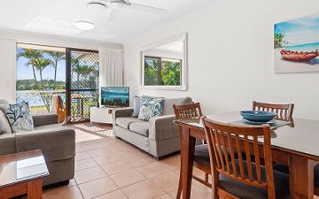 Riverside Apartment lounge area at Cayman Quays Noosaville, showcasing a large TV, dining furniture, and glass sliders transitioning out to a private patio with Noosa River views.
