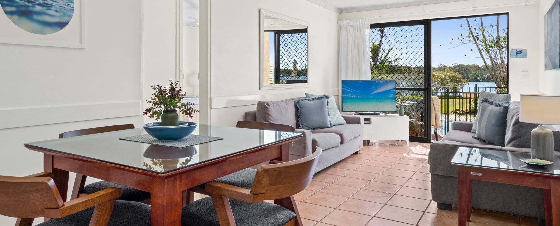 Fully self-contained holiday apartment interior at Cayman Quays featuring a modern lounge, big screen TV, dining area, and glass sliding doors opening to Noosa River balcony views.