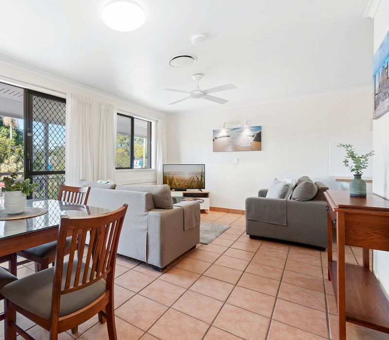 Larger open-plan living zone of a Standard Apartment featuring a lounge with a big screen TV, dining area, and glass sliders transitioning to a private balcony.