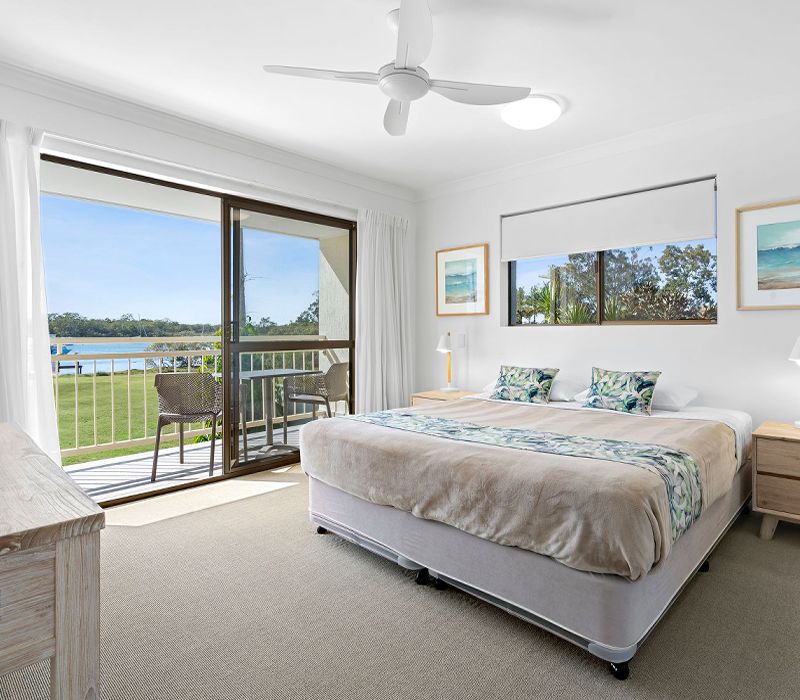 Luxury townhouse master bedroom retreat styled with a king-size bed and expansive glass sliders that open onto an elevated private balcony with panoramic Noosa River views.