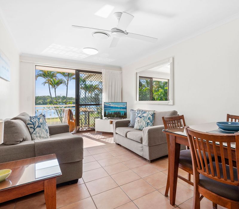 Comfortable self-contained lounge and dining area with a large flat-screen TV and glass sliders stepping directly onto a private ground-floor patio with Noosa River vistas.